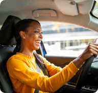 A woman in a yellow shirt drives a car and smiles broadly.